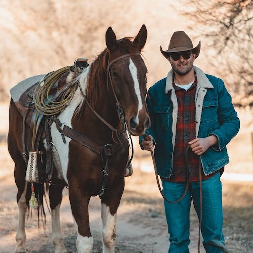 cowboy in jean jacket standing beside paint horse